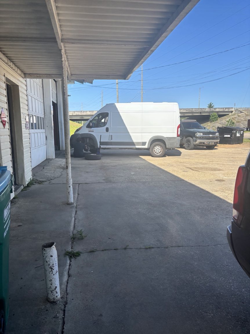 A J & P mechanic doing a wheel and brake job on a white cargo van under the shop awning, with the interstate overpass behind.